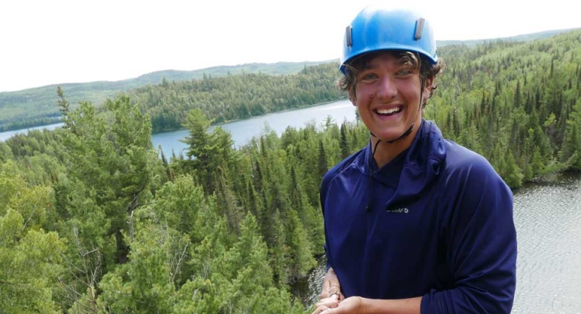 A person wearing a helmet smiles excitedly. They appear to be at high elevation. There is water and green trees below them. 